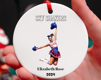 May include: White round ceramic ornament with a red ribbon. The ornament features a photo of a cheerleader in a blue and red uniform, jumping in the air with blue pom-poms. The text "SKY SHAKERS" is at the top of the ornament. Below the photo is the text "Elizabeth Rose 2024".