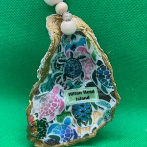 May include: A gold-trimmed oyster shell with a colorful design of sea turtles. The shell has a white string attached with wooden beads. The shell has a label that reads "Hilton Head Island".