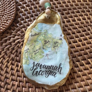 May include: An oyster shell ornament with a map of Savannah, Georgia, and the city's name in cursive. The shell has a gold-painted edge and is strung with a natural twine and wooden beads. The background is a woven basket.