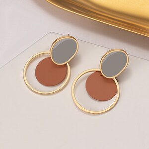 Può includere: Un paio di orecchini a cerchio dorati con un design circolare marrone e grigio. Gli orecchini sono in metallo e hanno uno stile semplice e moderno.