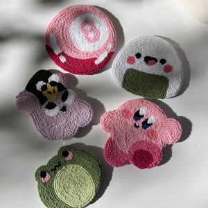 Pode incluir: Cinco  tapetes coloridos feitos à mão com vários personagens de desenhos animados. Os tapetes são feitos de fio e apresentam uma bola de 8 rosa, um sapo verde, um personagem rosa do Kirby, um onigiri branco e verde e um pinguim roxo.