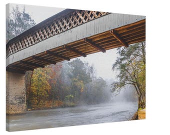 Può includere: Un ponte coperto in legno attraversa un fiume nebbioso in un ambiente boschivo. Il ponte è in legno con un tetto grigio e travi in legno. Il fiume scorre sotto il ponte ed è circondato da alberi. Gli alberi sono spogli e il terreno è ricoperto di foglie.
