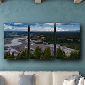 May include: A triptych landscape art piece depicting a river winding through a valley, with green trees and a blue sky. The artwork is displayed above a white sectional sofa. The dimensions are 36x72 inches / 91x182 cm.