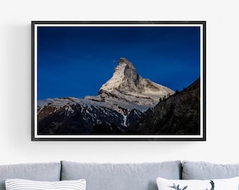May include: A framed photograph of a snow-capped mountain peak against a blue sky. The mountain is surrounded by trees and snow. The frame is black and the image is 24x36 inches.