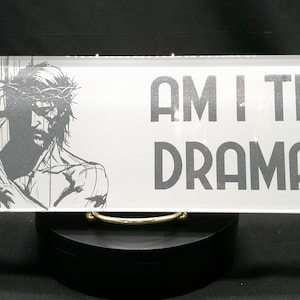 May include: A white glass plaque with a black and white image of a man carrying a cross. The text "AM I THE DRAMA?" is printed in black letters on the plaque.