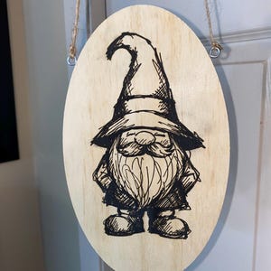 May include: Oval wooden sign featuring a hand-drawn illustration of a gnome with a long beard and pointed hat. The gnome is depicted in black ink on a light wood background. The sign is suspended by a natural twine cord.