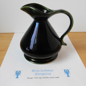 May include: A dark green ceramic jug with a wide base and a flared spout. The jug has a green glaze with a glossy finish. The handle is curved and has a green rim.