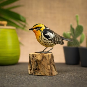 May include: A hand-carved wooden bird figurine with yellow, black, and orange plumage, perched on a small wooden stump. The bird has detailed feather markings and stands approximately 10 cm tall, showcasing a naturalistic design.
