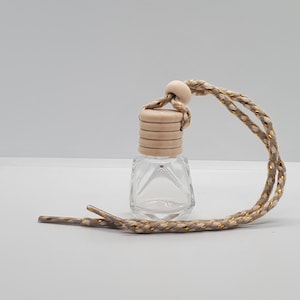 May include: A clear glass car air freshener bottle with a wooden cap and a braided beige and gold cord. The bottle has a geometric shape and is designed to hang in a vehicle. The cord is looped and knotted.