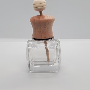 May include: A clear, square glass bottle with a wooden cap and a decorative wooden ball on top. The bottle has a reed diffuser stick inside. The wooden components are light brown. The bottle is designed for aromatherapy or fragrance use.