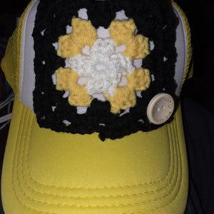 May include: A yellow baseball cap with a black, white, and yellow crocheted square design. The crocheted square features a floral pattern. A small, round button with the words "made with love" is attached to the cap.