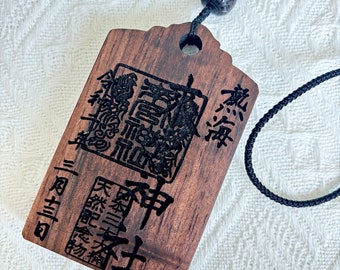 以下が含まれることがあります： 黒い紐の付いた、濃い茶色の木製の御守。御守には「天照大神」と「二柱神社」の文字が書かれている。