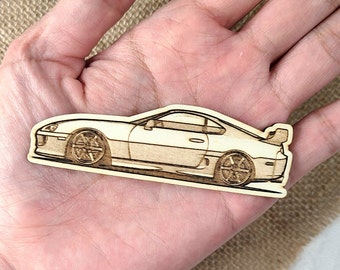 May include: A wooden cutout of a white Toyota Supra sports car. The car is in profile view and has black wheels.