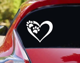 以下が含まれることがあります： 赤い車のリアウィンドウに貼られた、白いビニール製のステッカー。ハートの形をしていて、中に2つの肉球の跡があります。