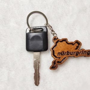 Könnte beinhalten: Ein silberner Schlüsselanhänger mit einem schwarzen Autoschlüssel und einem Holzanhänger in Form der Nürburgring-Rennstrecke mit dem eingravierten Wort "Nürburgring".