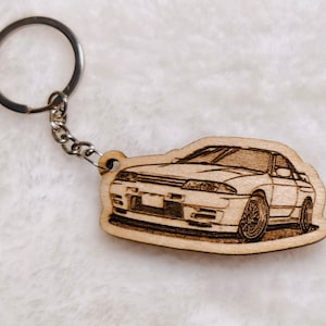May include: A wooden keychain featuring a detailed laser-engraved illustration of a white sports car. The keychain is attached to a silver metal ring.