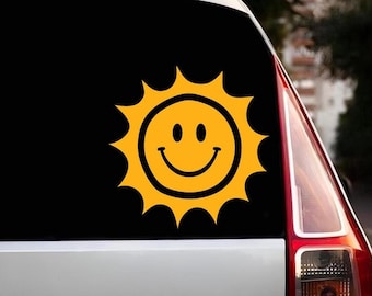 以下が含まれることがあります： 黒い車の黄色い笑顔の太陽のステッカー。太陽はシンプルで漫画のようなデザインです。