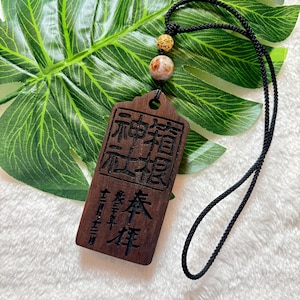 Puede incluir: Un colgante de madera con caracteres chinos tallados en él, colgando de un cordón negro con una cuenta blanca y dorada. El colgante es rectangular y tiene esquinas redondeadas. Los caracteres están en un estilo tradicional y son difíciles de leer.