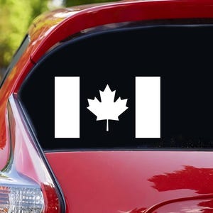 May include: A white Canadian flag decal with a red maple leaf in the center. The decal is applied to the back window of a red car.
