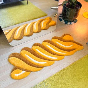 K&ouml;nnte beinhalten: Ein abstrakter Teppich in Gelb-, Braun- und Wei&szlig;t&ouml;nen. Der Teppich hat eine wellenf&ouml;rmige, l&auml;ngliche Form mit einer strukturierten Oberfl&auml;che. Das Design zeigt abwechselnde Streifen in Gelb, Braun und Wei&szlig;, wodurch ein ansprechendes Muster entsteht.