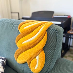 K&ouml;nnte beinhalten: Ein dekorativer, abstrakter Teppich in Gelb-, Braun- und Wei&szlig;t&ouml;nen liegt auf einem tealfarbenen Cordsofa. Der Teppich hat ein weiches, strukturiertes Aussehen und ein einzigartiges, flie&szlig;endes Design. Die Form des Teppichs erinnert an eine stilisierte Blume oder eine abstrakte Form.