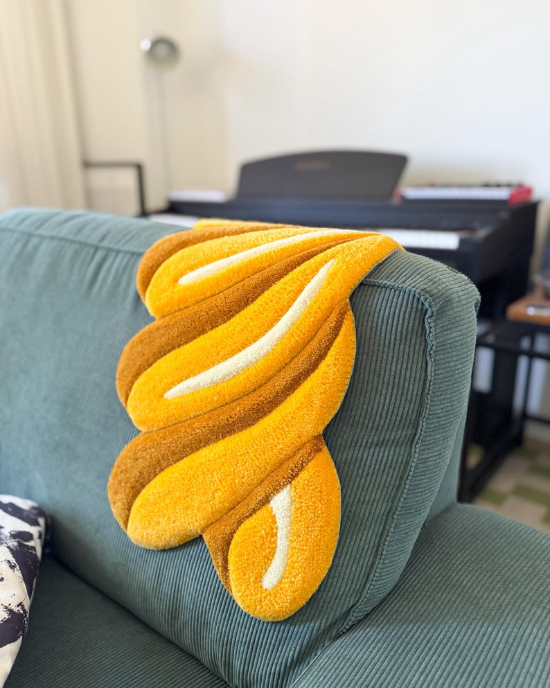 K&ouml;nnte beinhalten: Ein dekorativer, abstrakter Teppich in Gelb-, Braun- und Wei&szlig;t&ouml;nen liegt auf einem tealfarbenen Cordsofa. Der Teppich hat ein weiches, strukturiertes Aussehen und ein einzigartiges, flie&szlig;endes Design. Die Form des Teppichs erinnert an eine stilisierte Blume oder eine abstrakte Form.