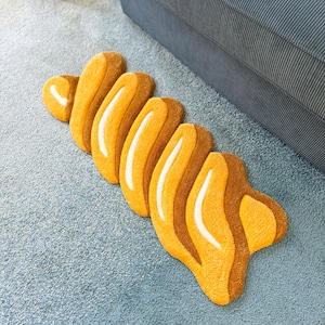 K&ouml;nnte beinhalten: Ein abstrakter Teppich in Gelb- und Braunt&ouml;nen, der einem Croissant oder einer stilisierten organischen Form &auml;hnelt. Der Teppich hat eine strukturierte Oberfl&auml;che und liegt auf einem hellblauen Teppich.