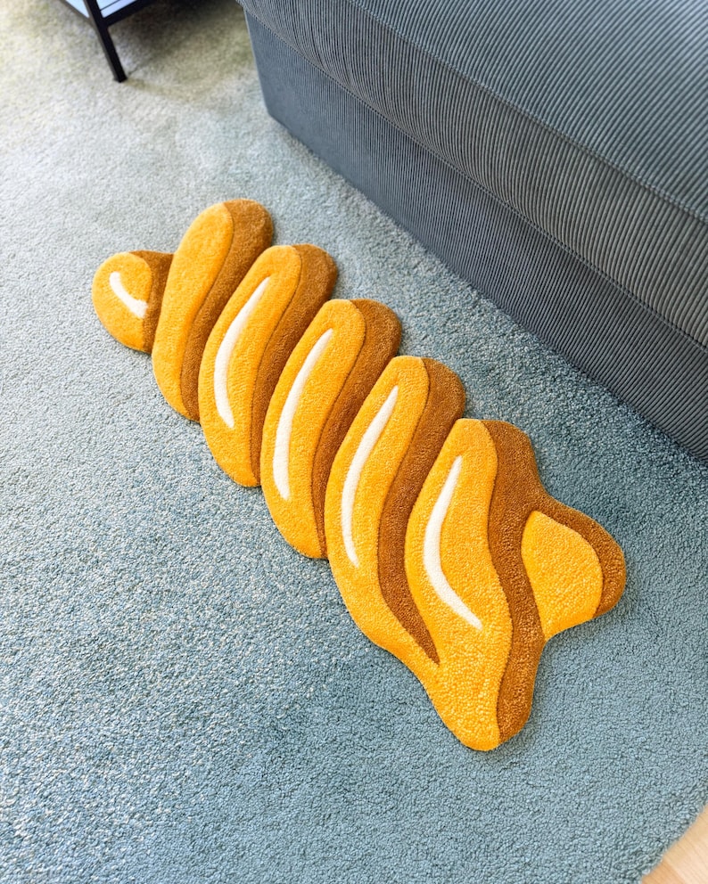 K&ouml;nnte beinhalten: Ein abstrakter Teppich in Gelb- und Braunt&ouml;nen, der einem Croissant oder einer stilisierten organischen Form &auml;hnelt. Der Teppich hat eine strukturierte Oberfl&auml;che und liegt auf einem hellblauen Teppich.