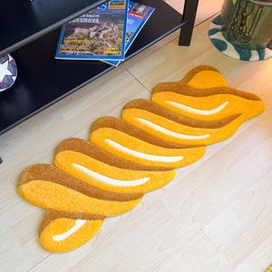 K&ouml;nnte beinhalten: Ein abstrakter Teppich in Gelb- und Braunt&ouml;nen mit einem wellenf&ouml;rmigen, geschichteten Design. Der Teppich liegt auf einem hellen Holzboden und verleiht der Raumdekoration einen Hauch moderner Kunst.