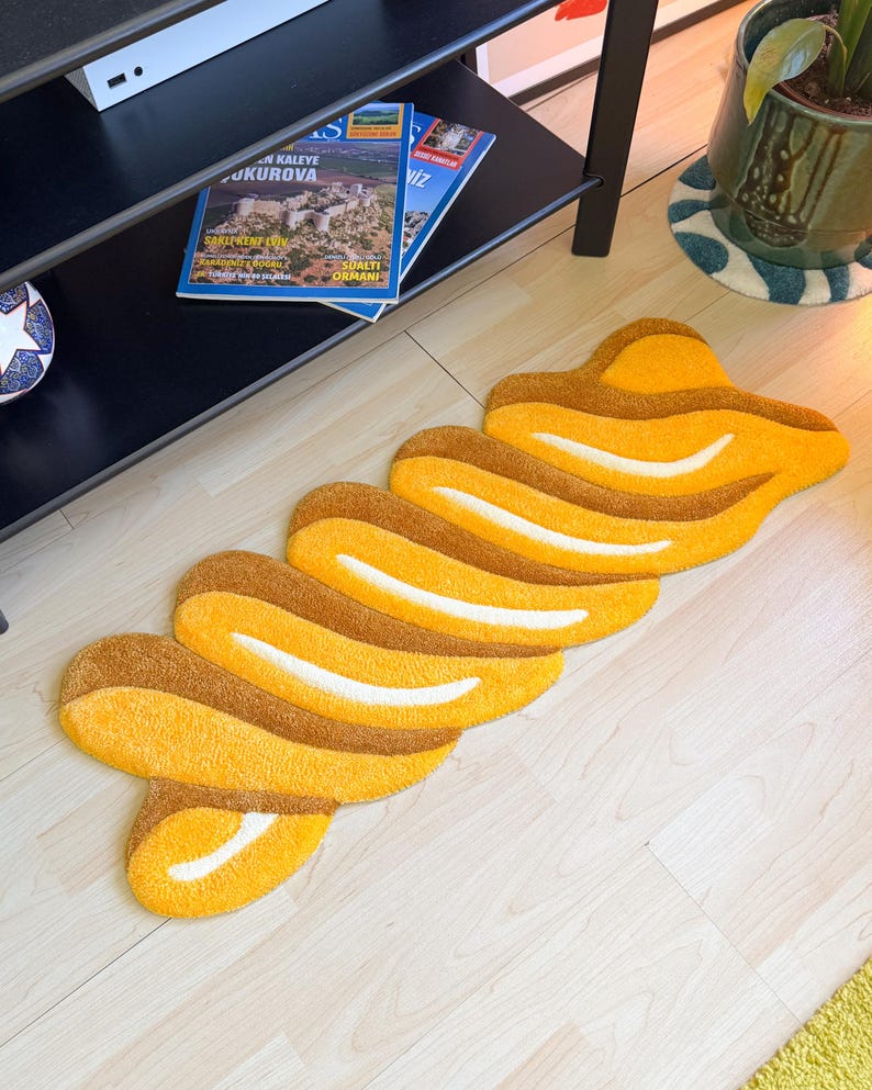 K&ouml;nnte beinhalten: Ein abstrakter Teppich in Gelb- und Braunt&ouml;nen mit einem wellenf&ouml;rmigen, geschichteten Design. Der Teppich liegt auf einem hellen Holzboden und verleiht der Raumdekoration einen Hauch moderner Kunst.