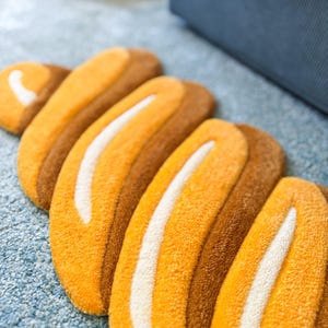 K&ouml;nnte beinhalten: Nahaufnahme eines Teppichs mit einzigartigem Design. Der Teppich zeigt eine Reihe von gebogenen, l&auml;nglichen Formen in Orange- und Braunt&ouml;nen, mit einem wei&szlig;en Streifen in der Mitte jeder Form. Der Teppich liegt auf einer hellblauen Oberfl&auml;che.