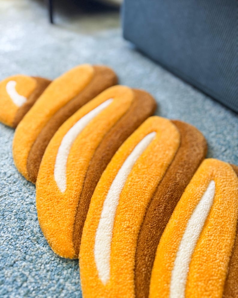K&ouml;nnte beinhalten: Nahaufnahme eines Teppichs mit einzigartigem Design. Der Teppich zeigt eine Reihe von gebogenen, l&auml;nglichen Formen in Orange- und Braunt&ouml;nen, mit einem wei&szlig;en Streifen in der Mitte jeder Form. Der Teppich liegt auf einer hellblauen Oberfl&auml;che.