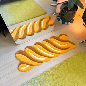 K&ouml;nnte beinhalten: Ein abstrakter Teppich in Wellenform mit braunen, gelben und wei&szlig;en Abschnitten. Das Design &auml;hnelt einem gro&szlig;en Geb&auml;ck. Der Teppich liegt auf einem hellen Holzboden, im Hintergrund ein gr&uuml;ner Teppich. Er ist etwa 150 cm lang.