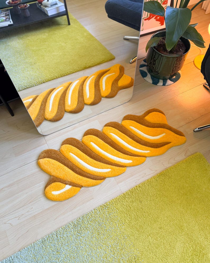 K&ouml;nnte beinhalten: Ein abstrakter Teppich in Wellenform mit braunen, gelben und wei&szlig;en Abschnitten. Das Design &auml;hnelt einem gro&szlig;en Geb&auml;ck. Der Teppich liegt auf einem hellen Holzboden, im Hintergrund ein gr&uuml;ner Teppich. Er ist etwa 150 cm lang.