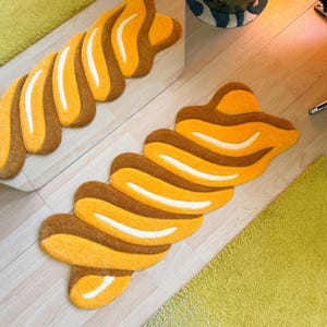 K&ouml;nnte beinhalten: Ein dekorativer Teppich in einer einzigartigen, abstrakten Form mit einem Muster aus abwechselnd braunen, gelben und wei&szlig;en Streifen. Der Teppich liegt auf einem hellen Holzboden, mit einem Spiegel, der das Design reflektiert. Im Hintergrund ist ein gr&uuml;ner Teppich zu sehen.