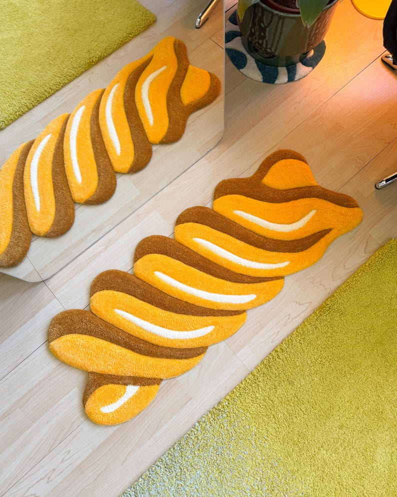 K&ouml;nnte beinhalten: Ein dekorativer Teppich in einer einzigartigen, abstrakten Form mit einem Muster aus abwechselnd braunen, gelben und wei&szlig;en Streifen. Der Teppich liegt auf einem hellen Holzboden, mit einem Spiegel, der das Design reflektiert. Im Hintergrund ist ein gr&uuml;ner Teppich zu sehen.