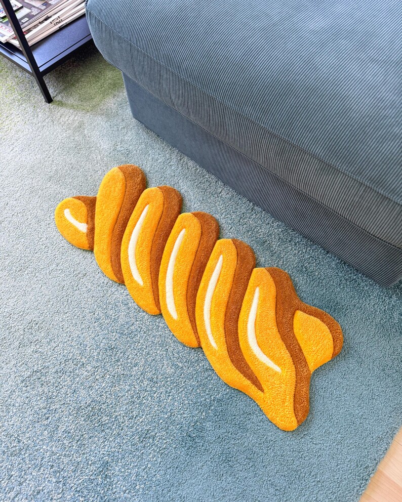 K&ouml;nnte beinhalten: Ein skurriler Teppich in Form eines Croissants, mit Orange- und Braunt&ouml;nen und wei&szlig;en Akzenten. Der Teppich liegt auf einem hellblauen Teppich, in der N&auml;he eines blauen Sofas.
