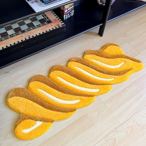 K&ouml;nnte beinhalten: Ein gelb-brauner Teppich mit einem einzigartigen, abstrakten Design. Der Teppich zeigt eine Reihe von miteinander verbundenen, geschwungenen Formen in Gelb- und Braunt&ouml;nen mit wei&szlig;en Akzenten. Er liegt auf einem hellen Holzboden.