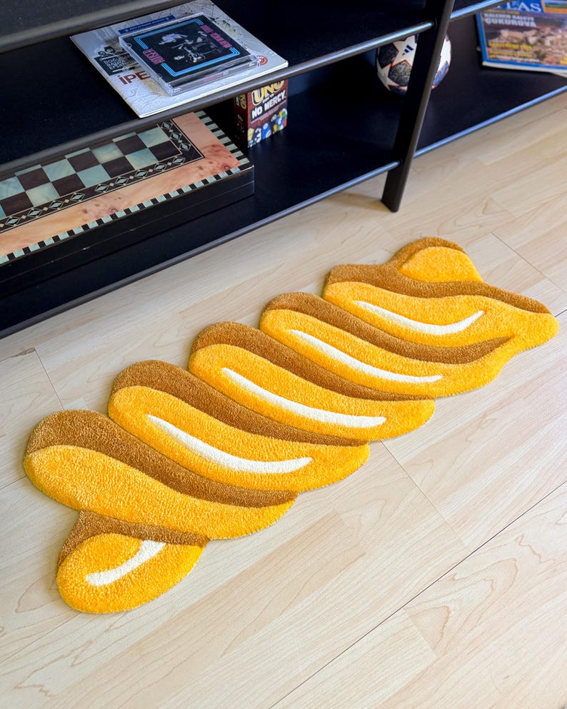 K&ouml;nnte beinhalten: Ein gelb-brauner Teppich mit einem einzigartigen, abstrakten Design. Der Teppich zeigt eine Reihe von miteinander verbundenen, geschwungenen Formen in Gelb- und Braunt&ouml;nen mit wei&szlig;en Akzenten. Er liegt auf einem hellen Holzboden.