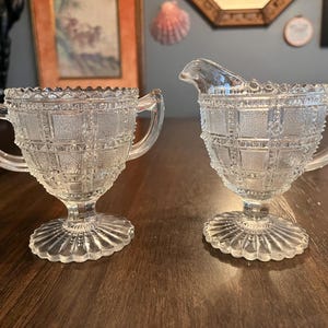 May include: Clear glass sugar bowl and creamer set. The sugar bowl has two handles and a decorative rim. The creamer has a spout and a handle. Both pieces have a textured, grid-like pattern and sit on a pedestal base.