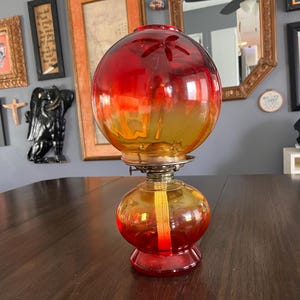 May include: A vintage oil lamp with a round, two-part glass body. The top globe is red and yellow, and the base is red. The lamp has a brass fitting and sits on a wooden table. Decorative home decor.
