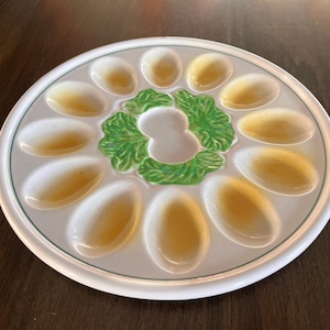May include: White ceramic deviled egg serving platter with 12 egg-shaped indentations and a green lettuce-shaped center. The eggs are filled with a yellow filling. The platter has a white rim with a green border.