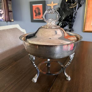 May include: A silver-plated serving dish with a matching lid featuring an ornate handle. The bowl is supported by three curved legs connected by a circular base. The polished metal surface reflects light, highlighting the smooth, rounded design.