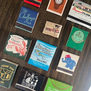 May include: A collection of vintage matchbooks with various designs and text, including logos, illustrations, and the names of businesses like restaurants and hotels. The matchbooks are arranged on a dark wooden surface.