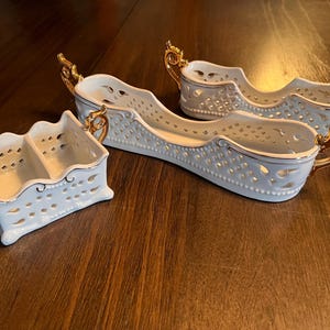 May include: A collection of three white ceramic serving dishes with gold-colored handles. The dishes have a pierced design and are arranged on a dark wooden surface. The serving dishes are in different shapes and sizes.