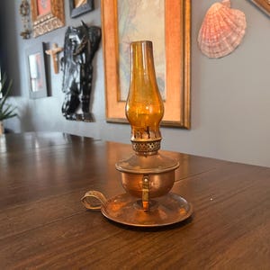 May include: An antique oil lamp with a copper base and a clear glass chimney. The lamp has a handle and a small round base. The glass chimney is amber-colored. The lamp is sitting on a dark wooden table.