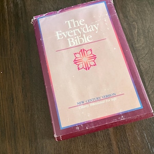 May include: A hardcover book titled "The Everyday Bible" with a pink and blue cover. The cover features a floral design and the text "New Century Version Clearly Translated For Life". The book is on a dark wood surface.