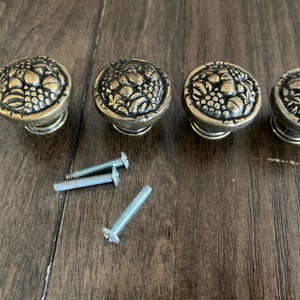 May include: Four round cabinet knobs with an antique brass finish, each featuring a raised, detailed relief pattern of assorted fruits including grapes and pears. Three silver-colored metal mounting screws are included with the hardware.