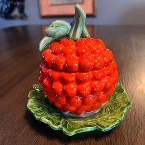 May include: A ceramic raspberry-shaped container with a lid, painted in a vibrant red hue. The container sits on a green leaf-shaped base. The lid features a green stem and leaves, adding to the realistic design.