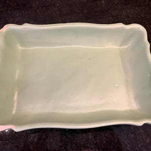 May include: A rectangular, light green ceramic dish with a scalloped edge. The dish has a smooth surface and a slightly irregular shape. The corners have a pinkish hue. This dish is suitable for serving food or as a decorative item.